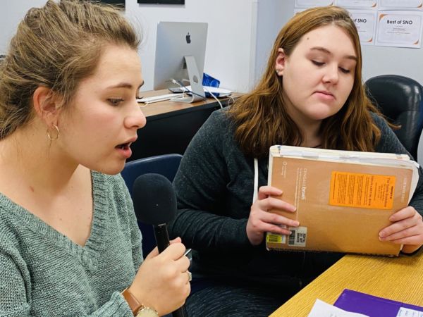 Rebecca Irby sings a parody written by Crusader staff for a special finals video. Cheyenne Miller waits for her part, which must be muffled behind a book.